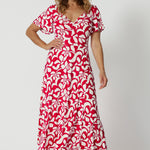 Over 40 year old woman's wears red and ivory coloured maxi floral dress. Dress for summer has a twist front bodice and flutter sleeves. Floral fabric used is easy-care and crease-resistant perfect for travel and easy packing. Leina & Fleur is a woman's clothing brand that actively stocks sizes from 8-24. Shop this summer ready dress in petite sizes and plus sizes.