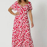 Over 40 year old woman's wears red and ivory coloured maxi floral dress. Dress for summer has a twist front bodice and flutter sleeves. Floral fabric used is easy-care and crease-resistant perfect for travel and easy packing. Leina & Fleur is a woman's clothing brand that actively stocks sizes from 8-24. Shop this summer ready dress in petite sizes and plus sizes.