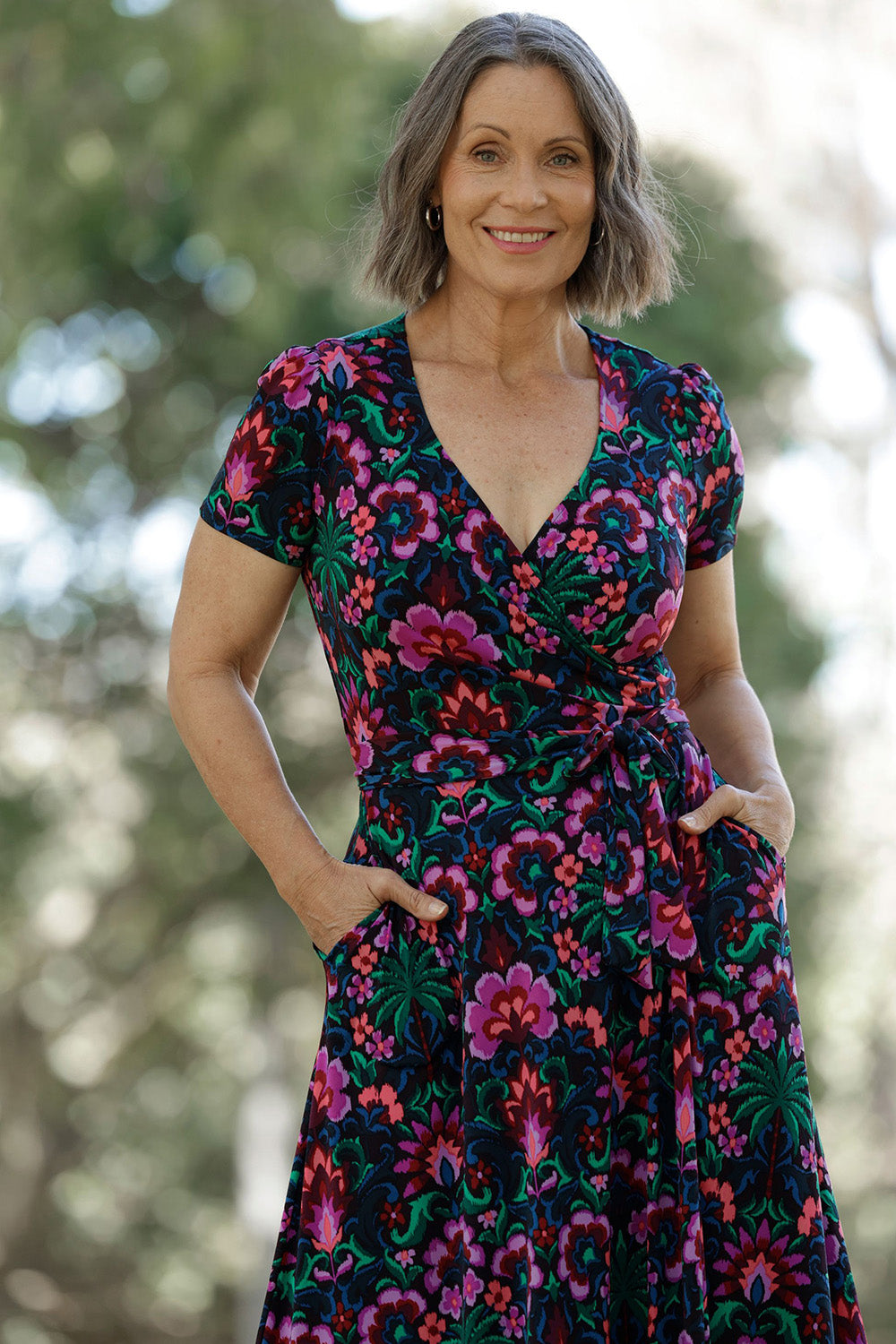 Featuring a striking floral print in bold shades of pink, green, burgundy, and blue, this dress flatters with its true wrap silhouette, extended waist ties, and a flowing A-line skirt.