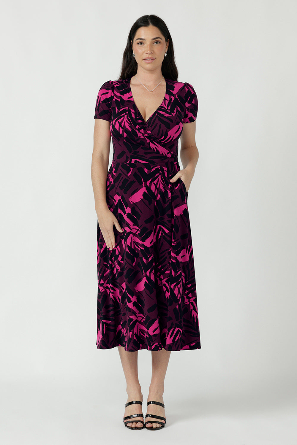 A woman modeling a short-sleeve midi wrap dress with a vibrant pink and black abstract pattern. Available in sizes 8-24.