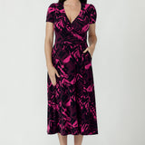 A woman modeling a short-sleeve midi wrap dress with a vibrant pink and black abstract pattern. Available in sizes 8-24.