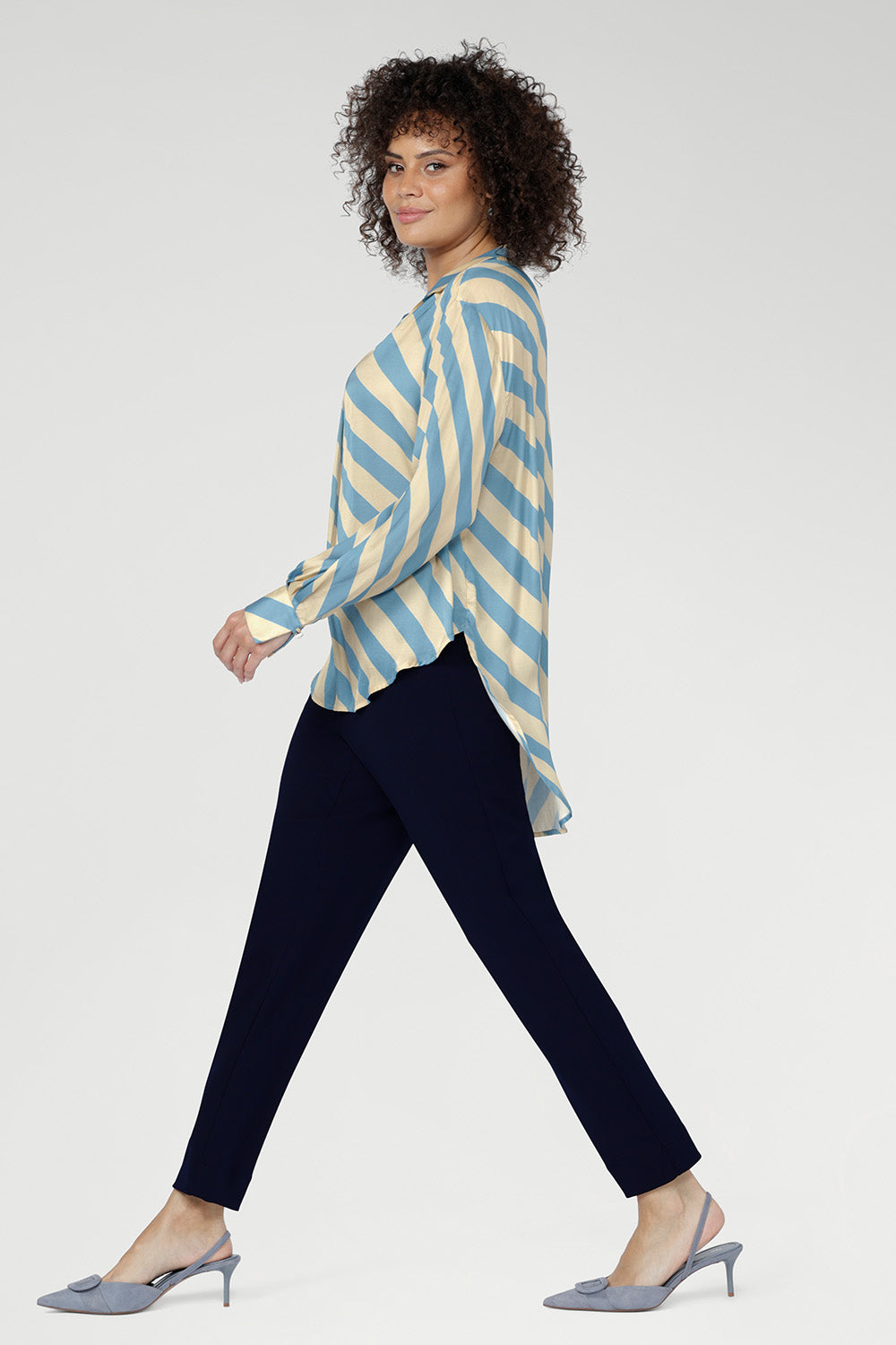 The Blue Sky Stripe blends soft sky blue and vanilla tones. Made in Australia. Women wears stripe blouse with tapered leg work pants in navy. 
