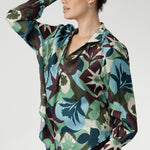 The bold abstract print blends rich tones of green, blue, and plum, making it a standout for both professional and social settings. A sophisticated collared V-neckline with tailored centre tuck adds structure, while the full-length cuffed sleeves with covered buttons bring a polished finish.