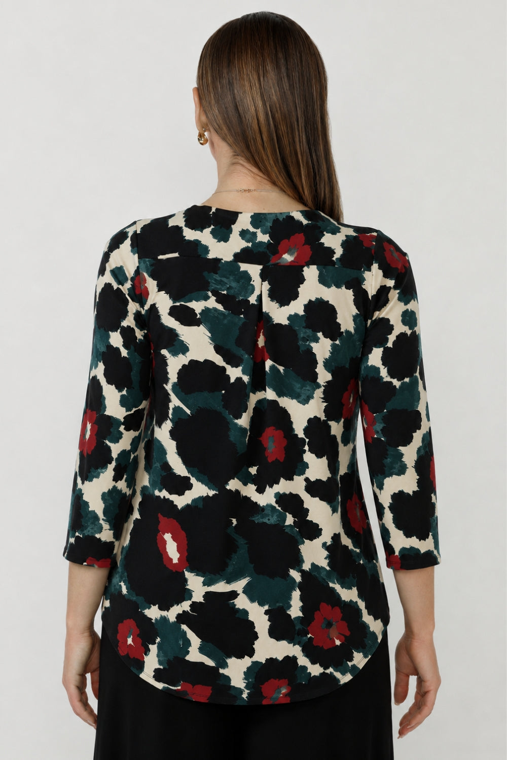 A woman viewed from the back wearing a three-quarter sleeve blouse with a bold abstract floral pattern in black, dark green, and red. Paired with black work pants. Australian-made designs in sizes 8-24.