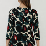A woman viewed from the back wearing a three-quarter sleeve blouse with a bold abstract floral pattern in black, dark green, and red. Paired with black work pants. Australian-made designs in sizes 8-24.
