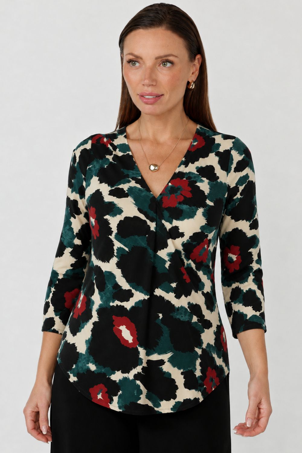 A woman wearing a three-quarter sleeve blouse with a bold abstract floral pattern in black, dark green, red, and cream, paired with black pants. Available in sizes 8-24.