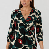 A woman wearing a three-quarter sleeve blouse with a bold abstract floral pattern in black, dark green, red, and cream, paired with black pants. Available in sizes 8-24.