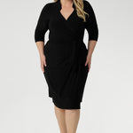 Plus size woman wearing a black wrap dress and black kitten heel. Tailored for a corporate or event setting and featuring a modern angular collar detail.
