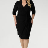 Plus size woman wearing a black wrap dress and black kitten heel. Tailored for a corporate or event setting and featuring a modern angular collar detail.