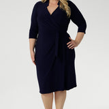 Plus size woman wearing a navy wrap dress and black kitten heel. Tailored for a corporate or event setting and featuring a modern angular collar detail.