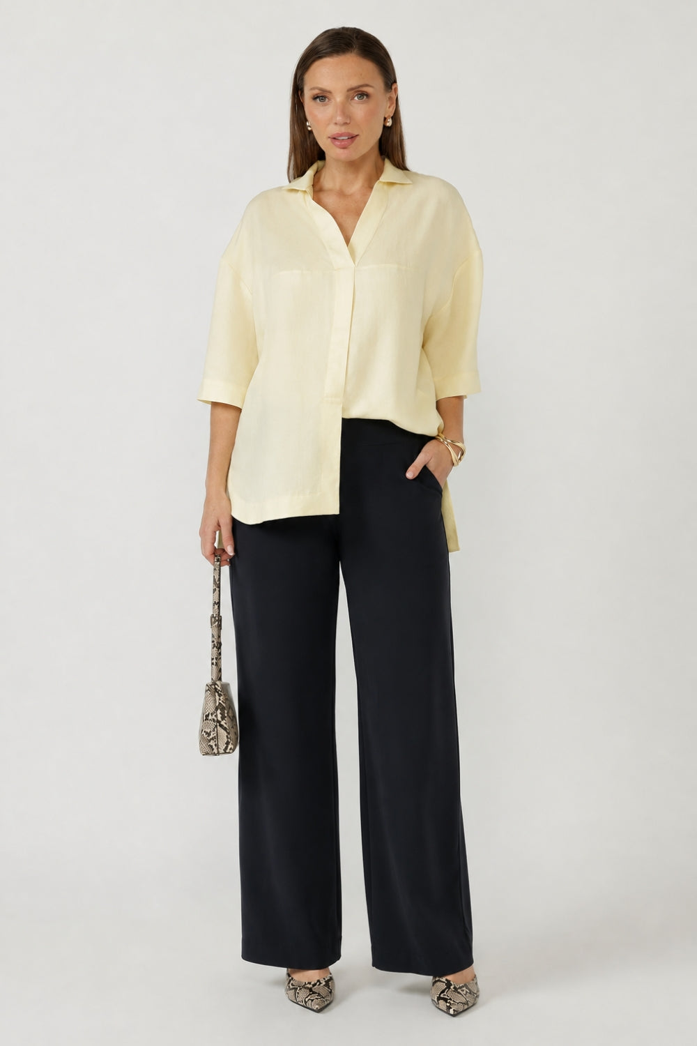 A woman modeling a pale yellow button-up shirt with three-quarter sleeves and dark wide-leg trousers, holding a small snakeskin-patterned handbag against a light gray studio background.