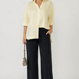A woman modeling a pale yellow button-up shirt with three-quarter sleeves and dark wide-leg trousers, holding a small snakeskin-patterned handbag against a light gray studio background.