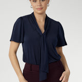 Close up shot of 30 year old woman wears navy work shirt with neck ties that can be styled in many different ways. This work shirt is perfect for a corporate look and pairs well with most Leina + Fleur core pieces. Easy care fabric is dry touch jersey. Garment is Australian made for women in Australia and New Zealand. Leina + Fleur is a women’s retail brand that proudly stocks sizes from 8-24. Shop this navy top in petite and plus sizes.