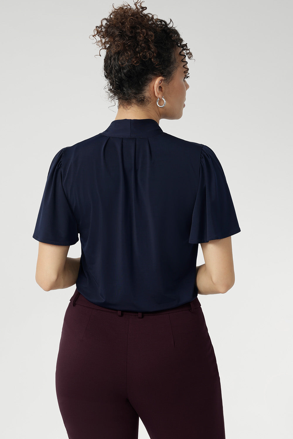 Close up image of Michael top from the back. 40 year old woman wears short sleeved work shirt with neck ties, tucked into work pants. Easy care dry touch jersey fabric used to create cool touch shirt. 