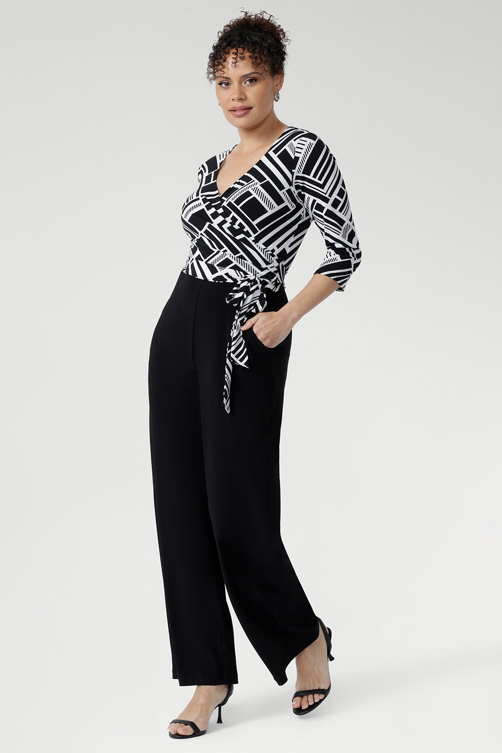 Showcasing a striking black and white geometric print across the wrap-style bodice, this jumpsuit features a flattering self-tie waist and a double-layered back for a refined and supportive fit. Available in sizes 8-24.