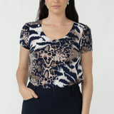 A woman modeling a short-sleeve scoop-neck top with an abstract animal-inspired print in navy, beige, and brown, paired with dark navy pants.