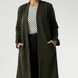 The Sorel Trench Coat is expertly tailored with a modern, sophisticated silhouette that elevates any wardrobe. Mature aged model wears winter Trenchcoat in Dark Olive over a olive stripe shirt and black work pants for women. 