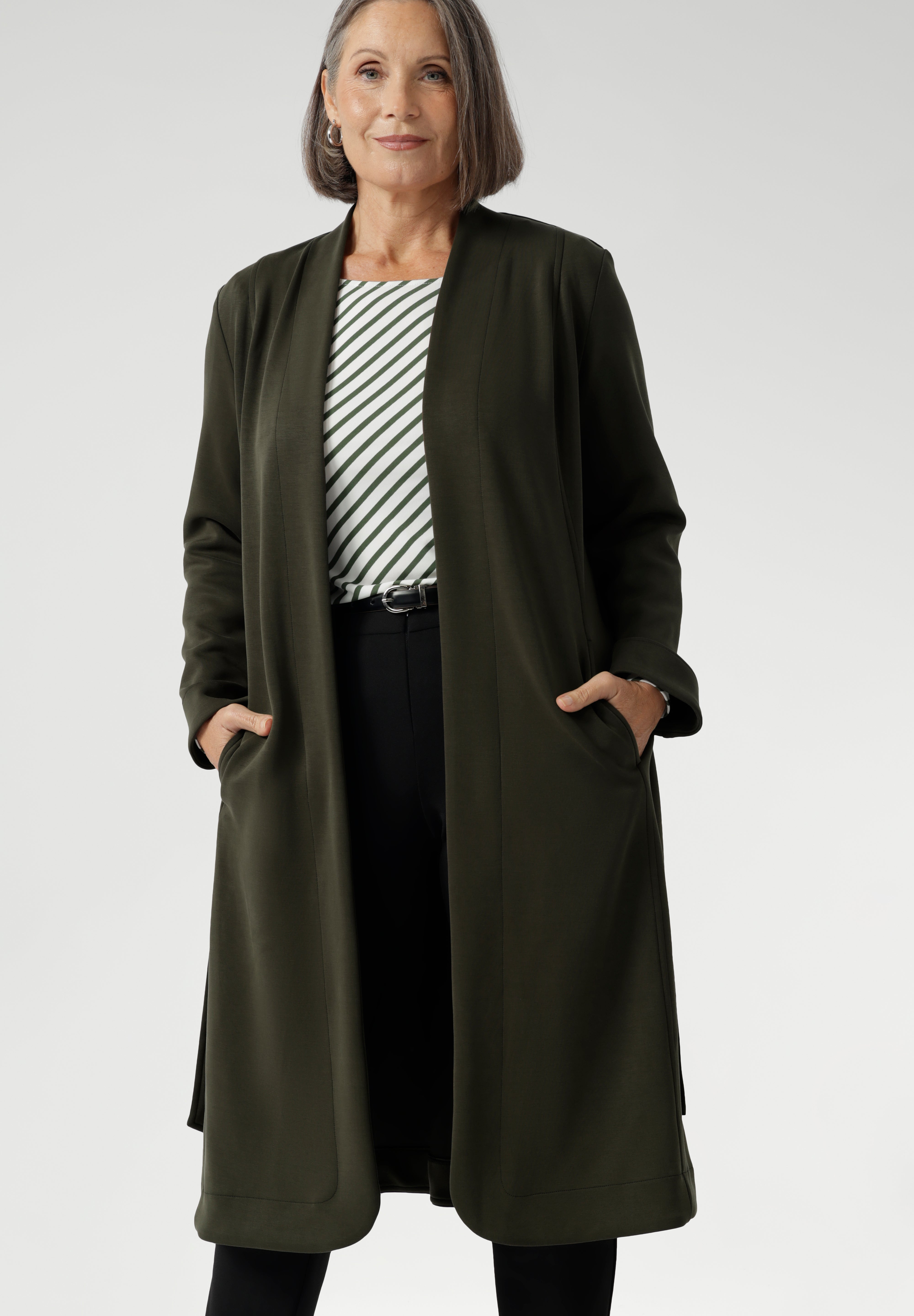 The Sorel Trench Coat is expertly tailored with a modern, sophisticated silhouette that elevates any wardrobe. Mature aged model wears winter Trenchcoat in Dark Olive over a olive stripe shirt and black work pants for women. 